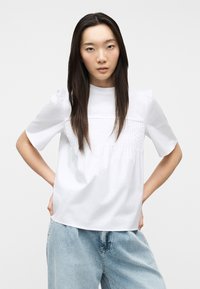 Νεαρή γυναίκα με μακριά σκούρα μαλλιά, φορώντας λευκό κοντομάνικο μπλουζάκι με λεπτομέρειες πιέτας και ανοιχτό μπλε τζιν, στέκεται με τα χέρια στη μέση.