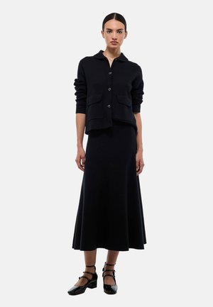 Cardigan en laine noir avec col, devant boutonné et deux poches, associé à une jupe longue noire assortie. Le mannequin porte des chaussures à brides noires.