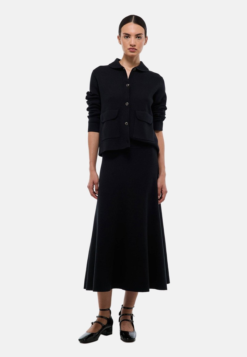 Cardigan en laine noir avec col, devant boutonné et deux poches, associé à une jupe longue noire assortie. Le mannequin porte des chaussures à brides noires.