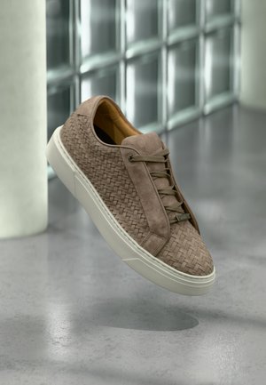 Brązowy pleciony skórzany sneaker z beżowymi sznurowadłami i białą podeszwą, pokazany unoszący się nad polerowaną betonową podłogą obok ściany z bloków szklanych.