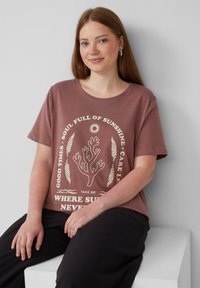 Braunes Baumwoll-T-Shirt mit rundem Halsausschnitt, mit weißen grafischen Text und einem korallenfarbigen Design. Kombiniert mit schwarzen Hosen, getragen von einer sitzenden Person.