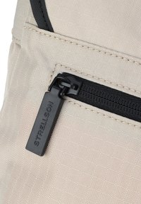 Zaino in tessuto beige con una tasca nera con zip e un tirazip rettangolare etichettato "STRELLSON". Superficie testurizzata con motivo a griglia.