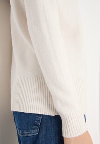 Weißer Strickpullover mit geripptem Bund und Ärmelabschlüssen. Weiche Textur, figurbetonte Silhouette und dezente Nähte. Kombiniert mit blauem Denim.