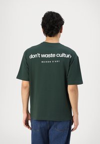 Zaļš kokvilnas T-krekls ar īsām piedurknēm. Mugurpusē ir lieli balti uzraksti "neiznieko kultūru" un "MAISON D'ART" zem tā.