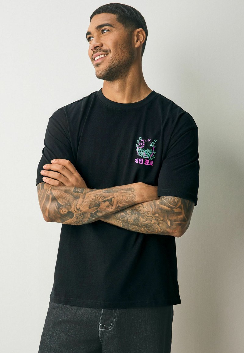 Tricou negru din bumbac, cu un design colorat imprimat pe piept, care prezintă text și grafică. Modelul are tatuaje vizibile pe brațe.