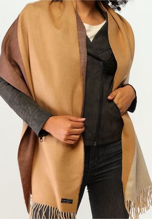 Personne portant un châle à franges beige et marron par-dessus une veste noire à fermeture éclair, les mains tenant le châle à l'avant.