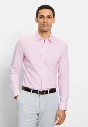 Mann trägt ein hellrosa Hemd mit Knöpfen, das in hellgraue Hose mit einem dunkelbraunen Gürtel gesteckt ist, und steht vor einem schlichten Hintergrund.