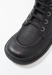 Botte en cuir noir avec un embout lisse, une surface texturée, des œillets en métal et des lacets noirs, avec des coutures contrastantes le long de la semelle.