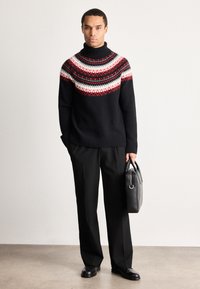 Homme portant un pull à col roulé noir à motifs, un pantalon noir, des chaussures noires, tenant une mallette en cuir noir, debout sur un sol en béton.