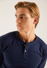 Cămașă henley albastru închis cu mâneci lungi, decolteu rotund și nasturi cu două capse. Țesătură moale cu textură netedă.