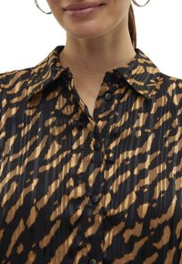 Blouse à motifs noirs et bruns avec un col, un boutonnage à l'avant et des plis verticaux, mettant en valeur une texture soyeuse.