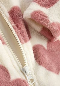 Fleece-Einteiler mit weißem Grund und großen, pinken Herzmustern. Verfügt über einen weißen Reißverschluss und einen weichen, strukturierten Stoff.