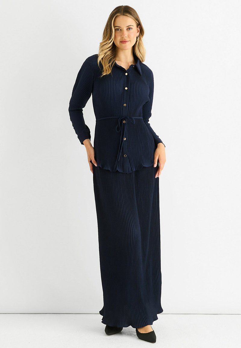 Woman standing in a navy blue ribbed long-sleeve buttoned top with waist tie and matching long skirt, wearing black pointed shoes.