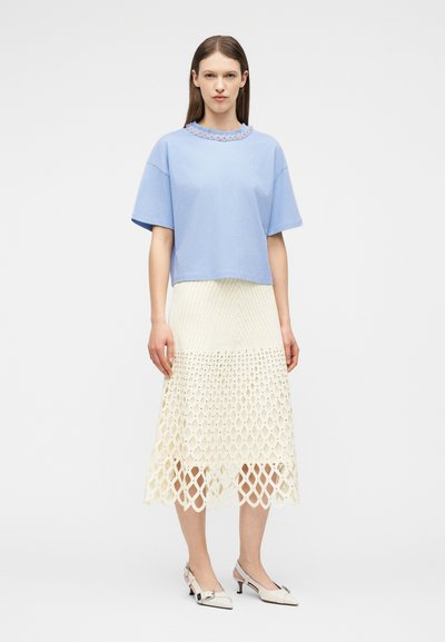 Γυναίκα που φοράει ένα ανοιχτό μπλε μπλουζάκι με κοντά μανίκια και διακόσμηση με πέρλες, καθώς και μια κρεμ μίντι φούστα με πλέγμα σχέδιο, συνδυασμένα με λευκά παπούτσια χαμηλού τακουνιού.