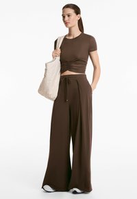 Top corto marrone con dettaglio arricciato abbinato a pantaloni larghi marroni e una borsa chiara. Tessuto morbido con una vestibilità rilassata.