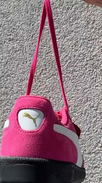 Sapatilha de camurça rosa com uma faixa branca acentuada, logótipo dourado no calcanhar e sola preta texturizada. Apresenta uma alça de tecido colorido.