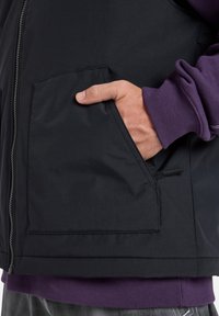 Giacca nera con zip frontale, dotata di tasca a pattina sul lato sinistro e polsini a coste viola a contrasto. Texture del tessuto liscia.