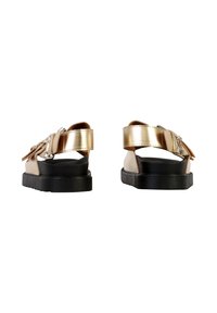 Paire de sandales ouvertes en métal doré avec semelles épaisses noires et brides à la cheville, vues de dos sur un fond blanc.