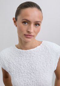 Weißes strukturiertes Oberteil mit rundem Ausschnitt und kurzen Ärmeln, das mit einem zerknitterten Stoff für zusätzliche Dimension und Details versehen ist.