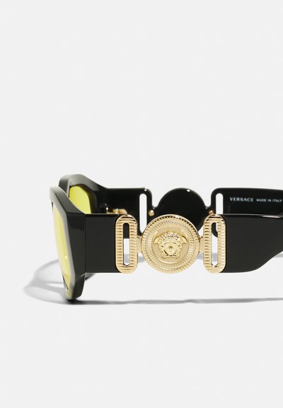 Óculos de sol retangulares pretos com lentes amarelas, apresentando um proeminente emblema em tom de ouro da Medusa de lado e detalhes em metal estriado.