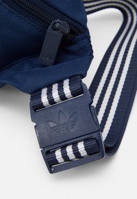 Mörkblå nylonväska rem med en plastspänne som har en upphöjd tre-strips logotyp och vävda ränder i vitt och mörkblått.