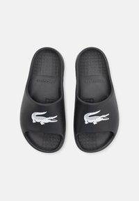Sandales noires en caoutchouc, avec une semelle texturée, une grande découpe pour le pied, et un logo crocodile blanc à l'avant.
