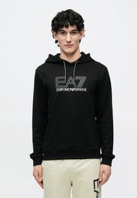 Čierna mikina s kapucňou z mäkkej látky s predným vreckom. Obsahuje biele logo EA7 Emporio Armani. Nastaviteľný šnúrka na kapucni.
