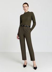 Femme debout avec une main dans la poche, portant un pull côtelé vert olive, un pantalon ajusté assorti, une ceinture noire et des stilettos noirs.