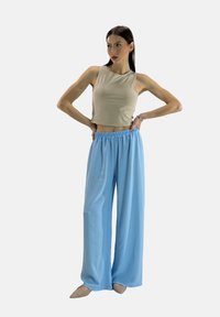Beige cropped tanktop gecombineerd met losse, lichtblauwe, gestructureerde wijde broek. Model in beige schoenen, staand in profiel.