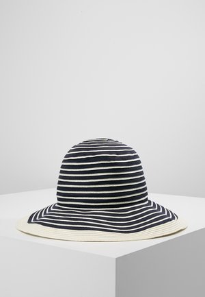 Chapeau de soleil à larges rayures bleu marine et blanc, avec un large bord, présenté sur un piédestal blanc sur un fond gris clair uni.