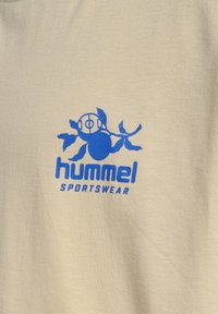 Beige bomulds t-shirt med et blåt "hummel" logo, der har en stiliseret figur og dekorative blade, som viser en glat tekstur.