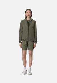 Giacca a zip verde oliva con cappuccio, due tasche frontali e dettagli neri, abbinata a pantaloni shorts coordinati. Modello che indossa scarpe da ginnastica bianche e calzini.