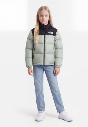 Junges Mädchen steht, trägt eine hellgrüne und schwarze Steppjacke, hellblaue Jeans und weiße Turnschuhe vor schlichtem Hintergrund.