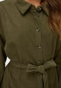 Robe chemise en velours côtelé vert olive avec un devant à boutons, un col et une ceinture nouée à la taille. Présente une texture côtelée subtile et des boutons noirs.