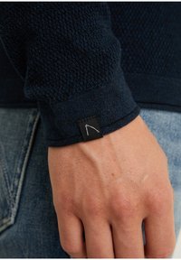 Granatowy sweter z dzianiny o fakturowanym wzorze, z czarną metką na rękawie. Widoczna dłoń, ukazująca teksturę skóry i detale.