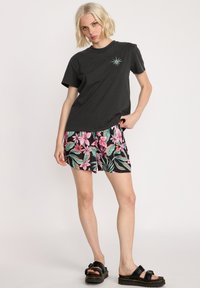 Czarny bawełniany t-shirt z turkusowym wzorem kompasu, w połączeniu z kwiecistymi szortami w kolorze czarnym, różowym i zielonym. Grube czarne sandały.
