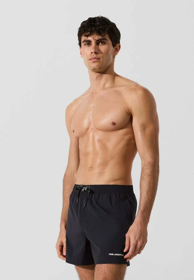 Homme musclé à la peau humide portant un short de bain noir Karl Lagerfeld, debout devant un fond gris uni.