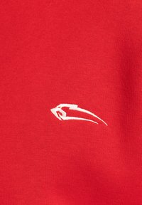 Tela de algodón roja con un pequeño logo bordado en blanco en una forma abstracta y estilizada, situado en la parte inferior izquierda.