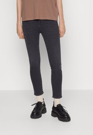Persoon die zwarte skinny jeans draagt, beige sokken, robuuste zwarte veterschoenen en een bruin topje, staand tegen een effen lichte achtergrond.