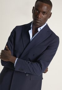 Blazer azul marino de corte cruzado con solapas de muesca, dos bolsillos frontales y puños abotonados. Se lleva sobre una camisa de color azul claro, con textura de tela suave.