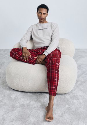 Sweatshirt gris avec le logo "TOMMY HILFIGER", pantalon à carreaux rouges, et pieds nus, assis sur une chaise sculpturale blanche texturée.