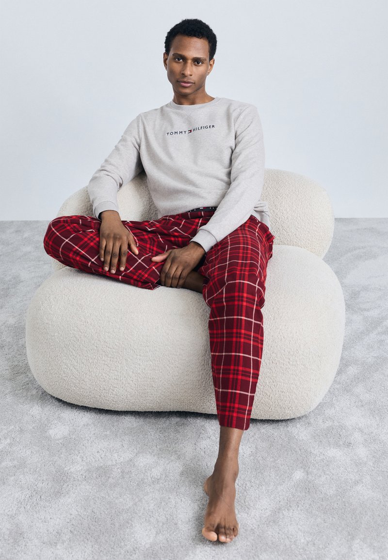 Hanorac gri cu logo-ul "TOMMY HILFIGER", pantaloni roșii cu linii, și picioare goale, așezat pe un scaun sculptural texturat de culoare albă.