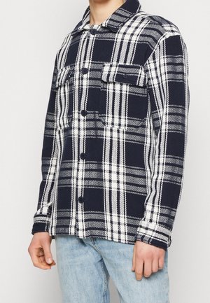Mann trägt ein marineblau-weiß kariertes Hemd mit Brusttaschen und hellblaue Jeans, steht vor einfachem Hintergrund.
