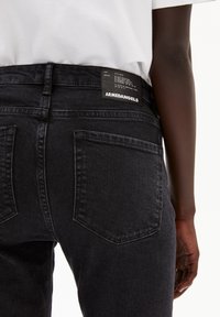 Jean en denim noir avec une taille haute et un design épuré. Comprend deux poches arrière et une étiquette sur la ceinture.