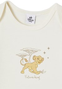 Cremfarbenes Baby-Body mit einer tanfarbigen Illustration eines Löwenjungen, begleitet von dem Text "Zukünftiger König" und Baumumrissen in einer passenden Farbe.