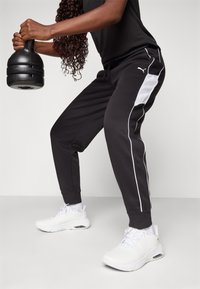 Calças de treino pretas com riscas brancas nas laterais, combinadas com tênis brancos. O indivíduo segura um kettlebell preto, exibindo uma pose ativa.