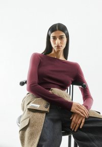 Bordeauxfarbenes Langarmshirt mit weitem Bootsausschnitt, kombiniert mit einem strukturierten braunen Wickel, über schwarzen Hosen mit weitem Bein. Leichter, glatter Stoff.