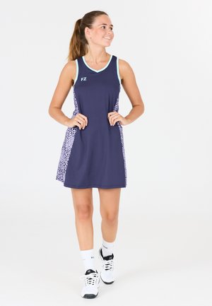 Jeune femme souriante, portant une robe de sport sans manches bleu marine avec des panneaux latéraux à motifs, chaussettes blanches et chaussures de sport blanches, debout sur fond blanc.