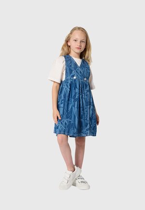 Jeune fille debout, portant une chemise blanche à manches courtes sous une robe sans manches bleue à motifs, avec des baskets blanches à scratch.