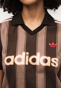 Καφέ και μπεζ ριγέ πόλο μπλουζάκι με μαύρο γιακά. Διαθέτει μεγάλο λογότυπο "adidas" σε ανοιχτό χρώμα και μικρό κόκκινο λογότυπο τριφυλλιού στα αριστερά.
